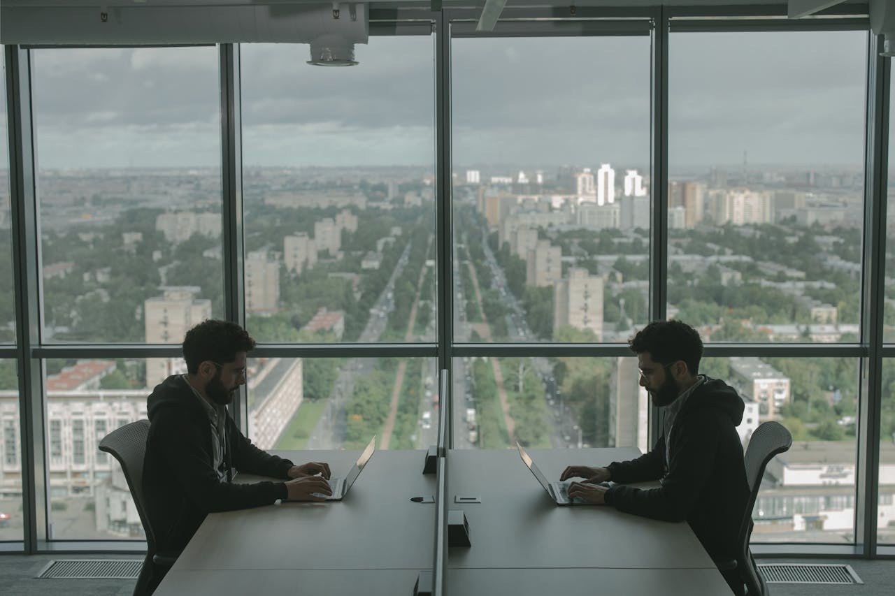 services-04 Two men working on laptops in a modern office with a panoramic city view through large windows.