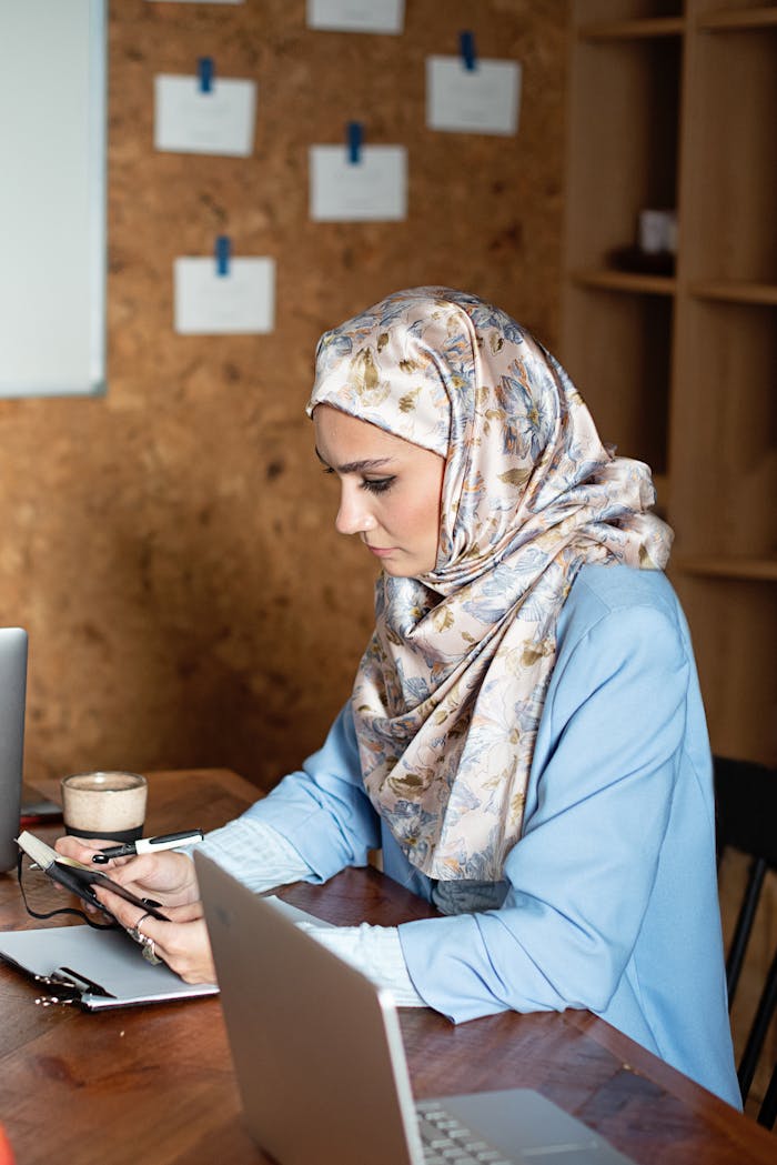 our-story A Muslim woman in hijab focuses on work at a wooden desk with laptops and a notebook in an office setting.